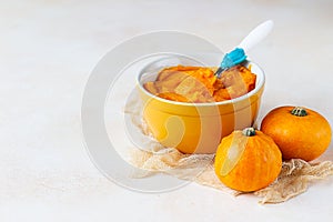 Homemade pumpkin puree in bowl with baby spoon and fresh pumpkins on light concrete background. The concept of baby food