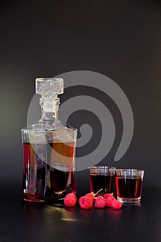 Homemade raspberry liqueur in two glasses and a bottle, ripe berries nearby on a black background