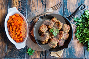 Homemade roasted beef meatballs in cast-iron skillet and beans baked in tomato sauce in baking dish on wooden table in kitchen