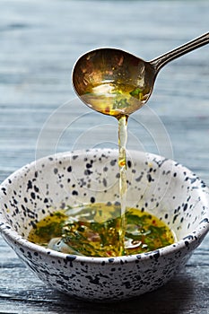 Homemade salad dressing with olive oil, vinegar, greens and spices pour from a spoon into a bowl on a gray wooden table.