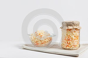 Homemade sauerkraut. Fermented food. Sauerkraut with carrots in glass jar and bowl with fork on wooden white background