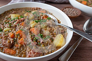 Lentil stew or soup with brown lentils, vegetables, potatoes and pork meat on a plate with spoon on wooden table.