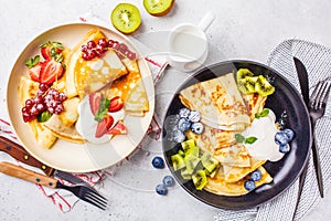 Homemade thin crepes served with curd cream, fruits and berries in black and white plates, top view