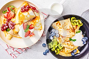 Homemade thin crepes served with curd cream, fruits and berries in black and white plates, top view