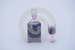 Homemade tincture of red cherry in a glass bottle and a wine crystal glass on a snow and white background