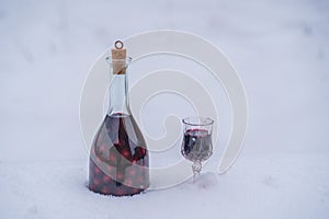 Homemade tincture of red cherry in a glass bottle and a wine crystal glass on a snow and white background