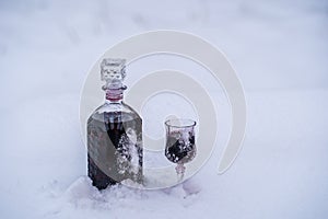 Homemade tincture of red cherry in a glass bottle and a wine crystal glass on a snow and white background