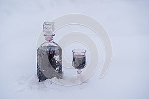 Homemade tincture of red cherry in a glass bottle and a wine crystal glass on a snow and white background