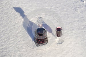 Homemade tincture of red cherry in a glass bottle and a wine crystal glass on a snow and white background