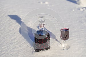 Homemade tincture of red cherry in a glass bottle and a wine crystal glass on a snow and white background