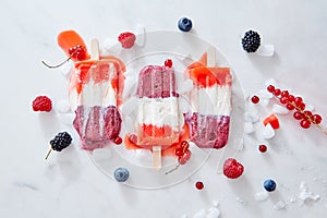 Tricolor strawberry and raspberry ice cream popsicles over ice with berries on marble background, top view and copy