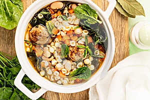 Homemade vegetable soup with meatballs and spinach in a white saucepan on a green background.