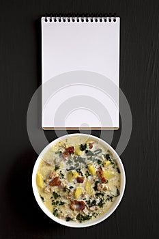 Homemade Zuppa Toscana with Kale and Bread in a white bowl, blank notepad on a black surface, top view. Overhead, from above, flat