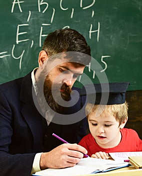 Homeschooling concept. Father checking homework, helps to boy, son. Teacher in formal wear and pupil in mortarboard in