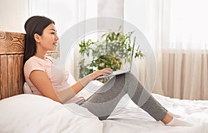 Homey Asian Woman Using Laptop Sitting In Bed At Home