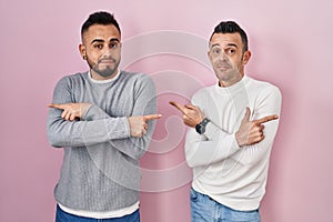 Homosexual couple standing over pink background pointing to both sides with fingers, different direction disagree