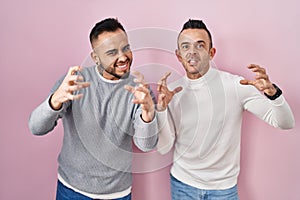 Homosexual couple standing over pink background shouting frustrated with rage, hands trying to strangle, yelling mad