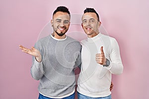 Homosexual couple standing over pink background showing palm hand and doing ok gesture with thumbs up, smiling happy and cheerful