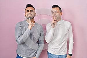 Homosexual couple standing over pink background thinking concentrated about doubt with finger on chin and looking up wondering