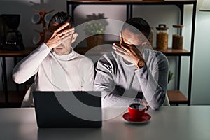 Homosexual couple using computer laptop covering eyes with hand, looking serious and sad