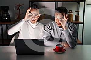 Homosexual couple using computer laptop doing ok gesture with hand smiling, eye looking through fingers with happy face