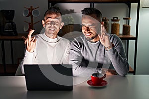 Homosexual couple using computer laptop gesturing finger crossed smiling with hope and eyes closed