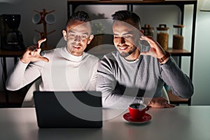 Homosexual couple using computer laptop smiling and confident gesturing with hand doing small size sign with fingers looking and