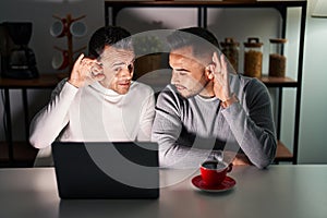 Homosexual couple using computer laptop smiling with hand over ear listening an hearing to rumor or gossip