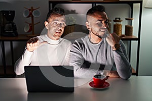 Homosexual couple using computer laptop smiling with happy face looking and pointing to the side with thumb up