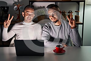 Homosexual couple using computer laptop smiling looking to the camera showing fingers doing victory sign