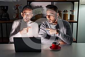 Homosexual couple using computer laptop surprised pointing with finger to the side, open mouth amazed expression