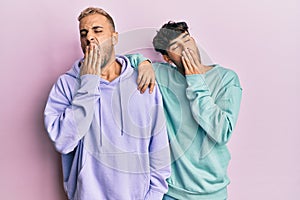 Homosexual gay couple standing together wearing casual clothes bored yawning tired covering mouth with hand