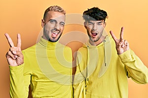 Homosexual gay couple standing together wearing yellow clothes smiling with happy face winking at the camera doing victory sign