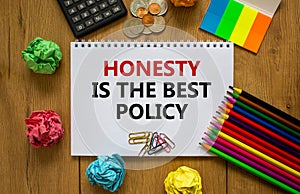 Honesty symbol. White note with words `honesty is the best policy` on beautiful wooden table, colored paper, colored pencils,