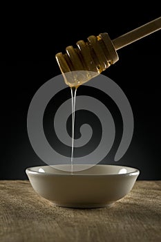 Honey dipper with honey dribling off into small white bowl on timber bench, low key