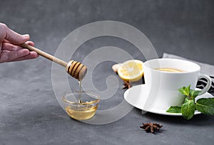 Honey flows from a wooden stick into a glass bowl close-up on a dark background with a cup of tea with lemon and mint