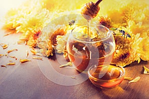 Honey in glass jar with bee flying and flowers on a wooden floor