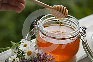 Honey in glass jar with bee flying and flowers on a wooden floor