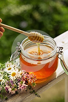 Honey in glass jar with bee flying and flowers on a wooden floor