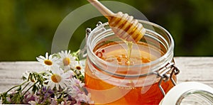 Honey in glass jar with bee flying and flowers on a wooden floor