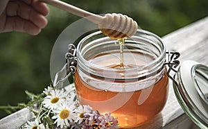 Honey in glass jar with bee flying and flowers on a wooden floor