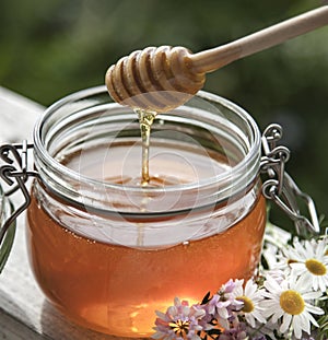 Honey in glass jar with bee flying and flowers on a wooden floor