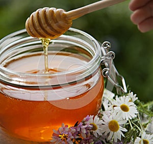 Honey in glass jar with bee flying and flowers on a wooden floor