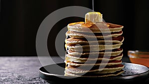 Honey or maple syrup is poured over a stack of pancakes with butter on dark background. Close-up high quality 4k footage