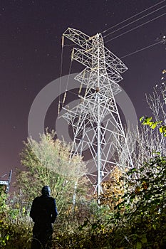 A hooded figure, back to camera. Looking up at a electricty pylon on a winters night