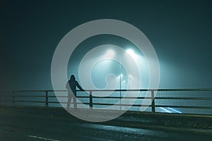 A hooded figure, standing with back to camera on a bridge, looking out at street lights. On a foggy night