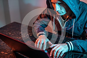 Hooded hacker is typing on a laptop keyboard in a dark room under a neon light. Cybercrime fraud and identity theft