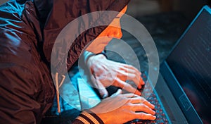 Hooded hacker is typing on a laptop keyboard in a dark room under a neon light. Cybercrime fraud and identity theft