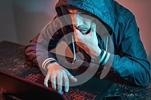 Hooded hacker is typing on a laptop keyboard in a dark room under a neon light. Cybercrime fraud and identity theft