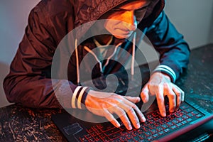 Hooded hacker is typing on a laptop keyboard in a dark room under a neon light. Cybercrime fraud and identity theft